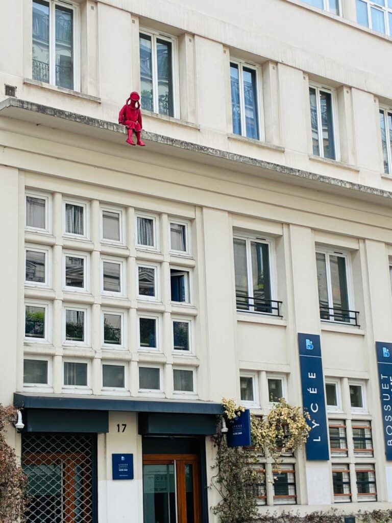 Red sculpture of a child sitting on a top-storey window ledge, looking down over the streets of Montmartre